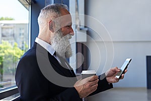 Smiling old man drinking coffee and surfing in phone