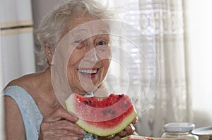 Smiling old senior woman holding a slice of fresh watermelon looking at camera at home background - healthy eating