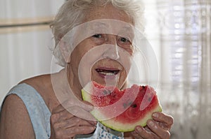 Smiling old senior woman holding a slice of fresh watermelon looking at camera at home background - healthy eating