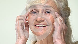 Smiling old woman stretching eye wrinkles up, face contouring anti-aging massage