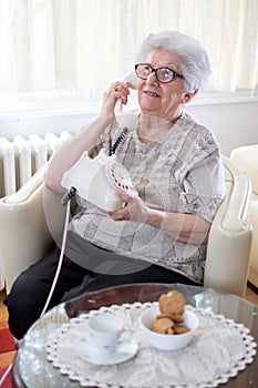 Smiling old woman talking on landline rotary phone while relaxing on armchair at home.