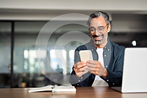 Smiling older Indian business man sitting at desk using mobile phone.