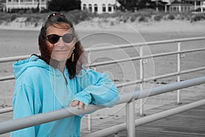 Smiling older woman in color with her arm on a boardwalk fence rail with the beach and fence in black and white