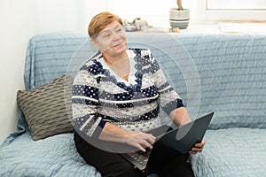 A smiling older woman at home is sitting on the couch with a laptop. Lifestyle. Positive emotions