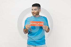 Smiling optimistic man holds card with subscribe inscription looking at camera with happy expression