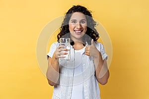 Smiling optimistic woman standing with glass of water, showing thumb up, like beverage.