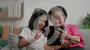 Smiling overjoyed mature grandmother with little granddaughter using tablet on cozy couch. Happy mature granny and small