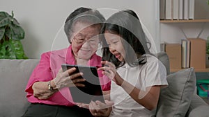 Smiling overjoyed mature grandmother with little granddaughter using tablet on cozy couch. Happy mature granny and small