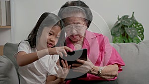 Smiling overjoyed mature grandmother with little granddaughter using tablet on cozy couch. Happy mature granny and small