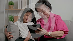 Smiling overjoyed mature grandmother with little granddaughter using tablet on cozy couch. Happy mature granny and small