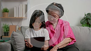 Smiling overjoyed mature grandmother with little granddaughter using tablet on cozy couch. Happy mature granny and small