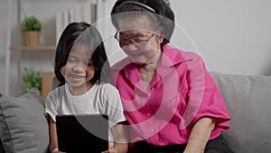 Smiling overjoyed mature grandmother with little granddaughter using tablet on cozy couch. Happy mature granny and small