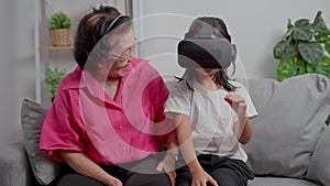 Smiling overjoyed mature grandmother with little granddaughter using VR on cozy couch. Happy mature granny and small grandchild