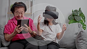 Smiling overjoyed mature grandmother with little granddaughter using VR on cozy couch. Happy mature granny and small grandchild