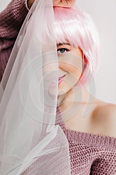 Smiling pale woman with brown eyes hiding face behind white cloth. Indoor close-up portrait of laughing girl in pink