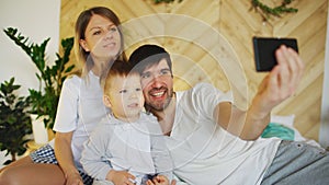 Smiling parents with baby taking selfie family photo on bed at home