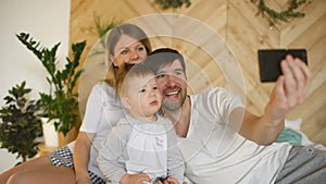 Smiling parents with baby taking selfie family photo on bed at home
