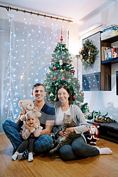 Smiling parents with a little girl and a cat on their knees sit near the Christmas tree on the floor