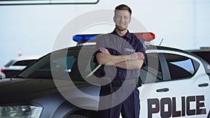 Smiling patrolman standing near car with crossed hands, carrying out duties