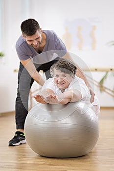 Smiling physiotherapy student helping senior woman lay on the exercising ball during rehabilitation