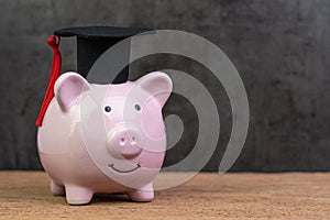 Smiling pink piggy bank wearing graduated hat on wooden table with dark black background and copy space, education fund,