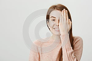 Smiling playful caucasian redhead with cute freckles hiding half of face behind palm, smiling cheerfully and standing