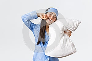 Smiling pleased cute asian girl in blue pyjama and sleeping mask, hugging pillow and stretching hands delighted as