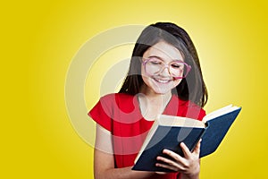 Smiling Portrait of a cute little schoolgirl loving to learn, holding with hands a book and wearing glasses