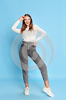 Smiling. Portrait of young beautiful girl in white blouse and jeans posing over blue studio background. Concept of