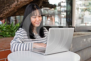 A smiling and positive Asian woman is working remotely outdoors in a city park, using her laptop