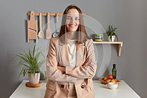 Smiling positive business woman with brown hair wearing beige jacket standing in home kitchen interior looking at camera keeps