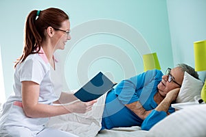 Smiling positive nurse reading a book to her old patient