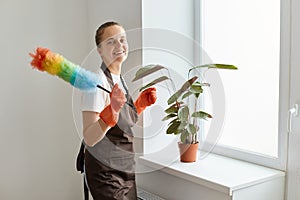 Smiling positive optimistic housewife wearing white t shirt and apron posing with ppduster in hands new windowsill with flower in