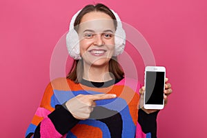 Smiling positive woman wearing jumper and fur earmuffs posing isolated over pink background, pointing at smart phone empty screen