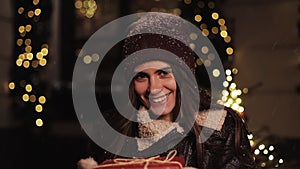 Smiling Preety Girl in Winter Hat and Mittens Holding a red Present Box, Looking Happy. Woman Standing at Lights Looking