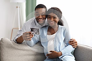 Smiling pregnant black couple holding ultrasound image, hugging