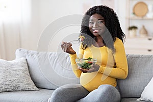 Smiling pregnant black woman eating fresh salad, sitting on sofa