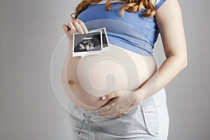 Smiling pregnant cute young caucasian woman standing against grey studio background and holding an ultrasound black and white scan