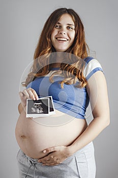 Smiling pregnant cute young caucasian woman standing against grey studio background and holding an ultrasound black and white scan