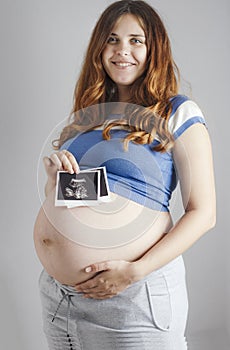 Smiling pregnant cute young caucasian woman standing against grey studio background and holding an ultrasound black and white scan