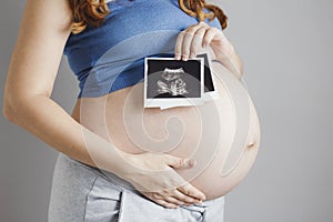 Smiling pregnant cute young caucasian woman standing against grey studio background and holding an ultrasound black and white scan
