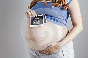 Smiling pregnant cute young caucasian woman standing against grey studio background and holding an ultrasound black and white scan