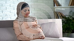 Smiling pregnant lady in hijab holding tummy, baby expectation, motherhood