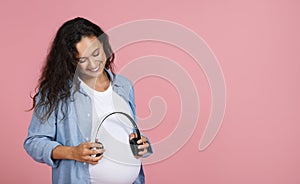 Smiling Pregnant Lady Standing Holding Headphones Near Belly, Pink Background