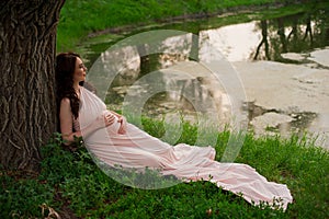 Smiling pregnant woman 25-29 year old resting by the lake. Posing outdoors. Motherhood. Maternity.