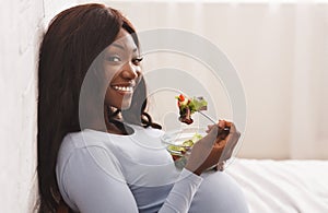 Smiling pregnant woman eating fresh salad on bed at home