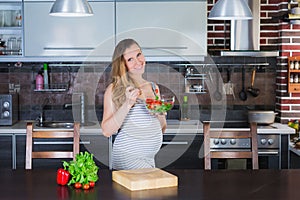 The smiling pregnant woman in kitchen is eating vegetable salad.