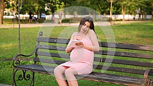 Smiling Pregnant woman sitting on bench in summer city park and talking on her mobile phone and making notes in notebook. Expecta