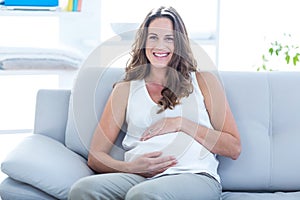 Smiling pregnant woman sitting with hand on stomach on sofa