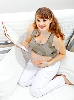 Smiling pregnant woman sitting on sofa with book.